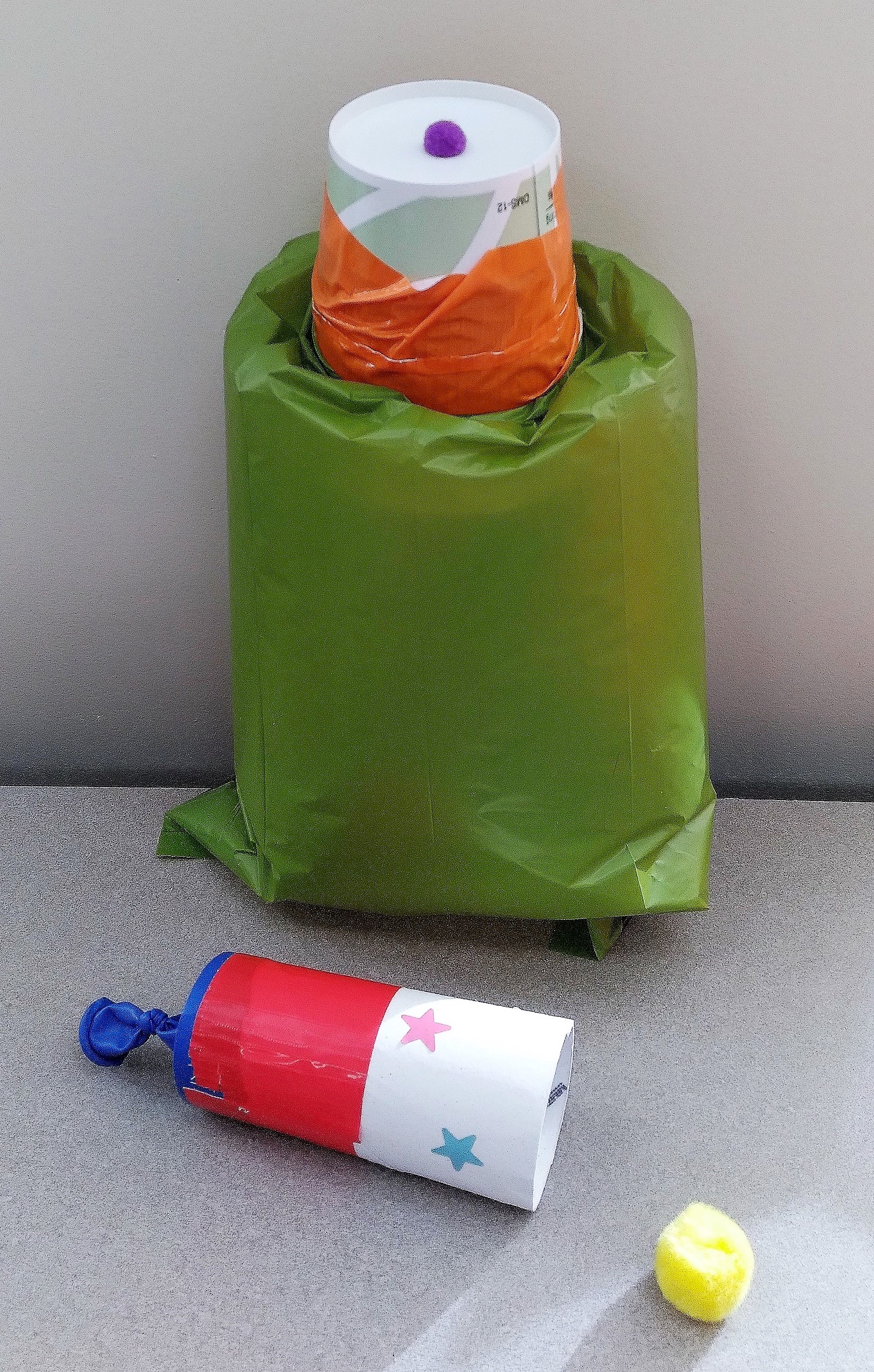 two contraptions, one with a green air filled bag and attached cup and purple pom pom and the second a white tube with blue balloon on the end and yellow pom pom