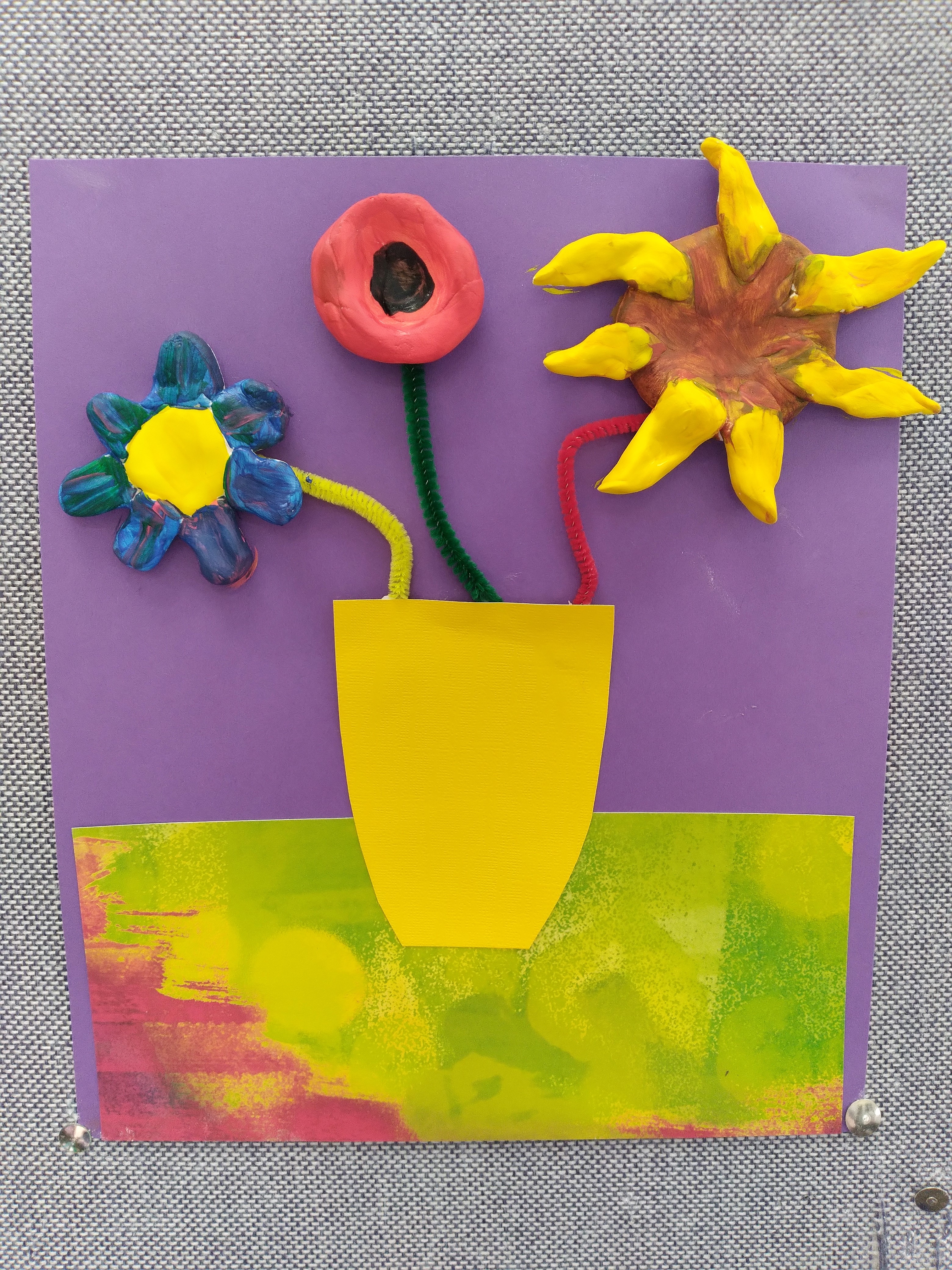 three clay flowers in a yellow paper vase on a green and purple background